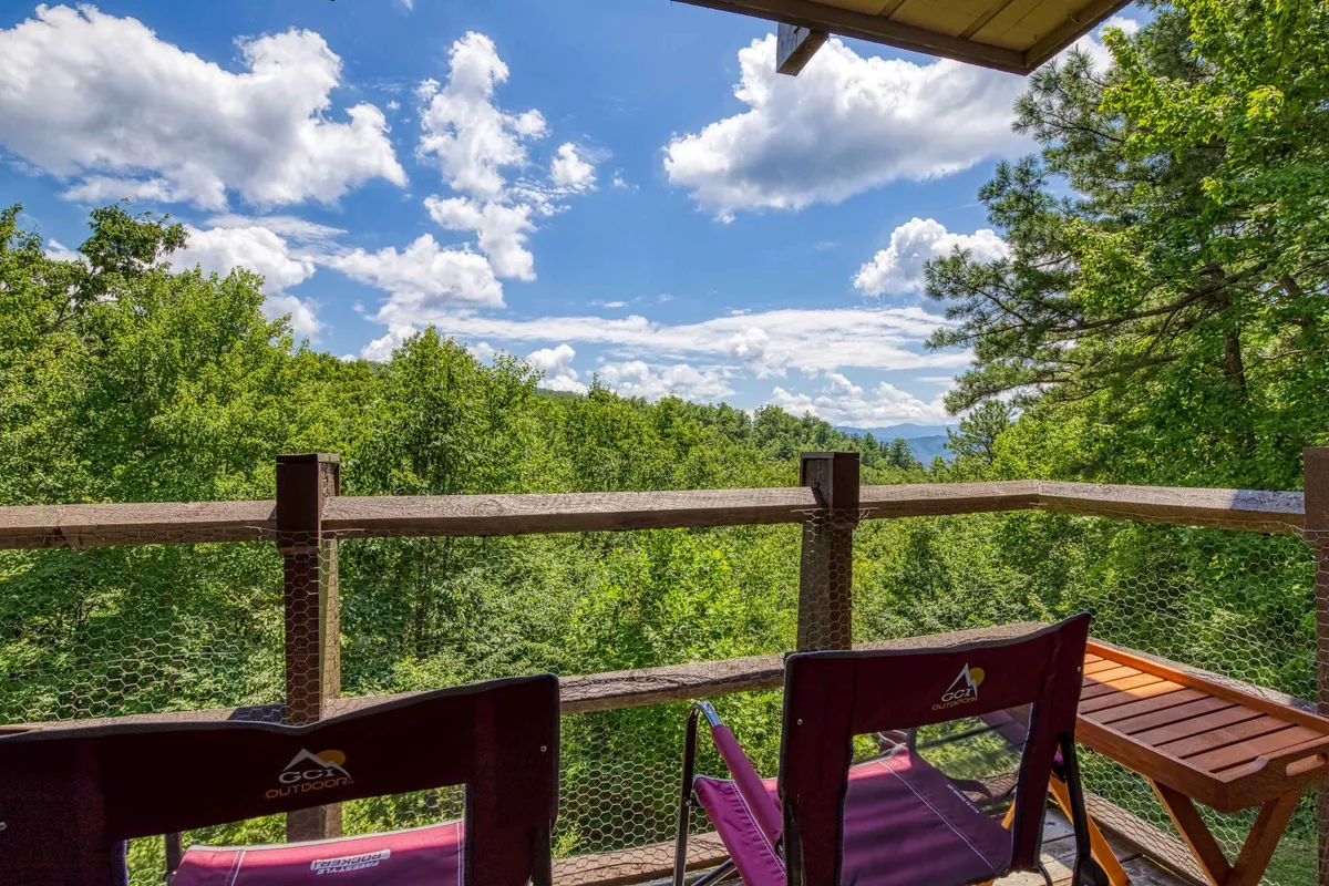 A private balcony is outfitted with two outdoor chairs and a sturdy deck railing, overlooking a scenic expanse of trees and distant mountains beneath a cloudy blue sky. The setup is designed for relaxation and taking in beautiful natural views.