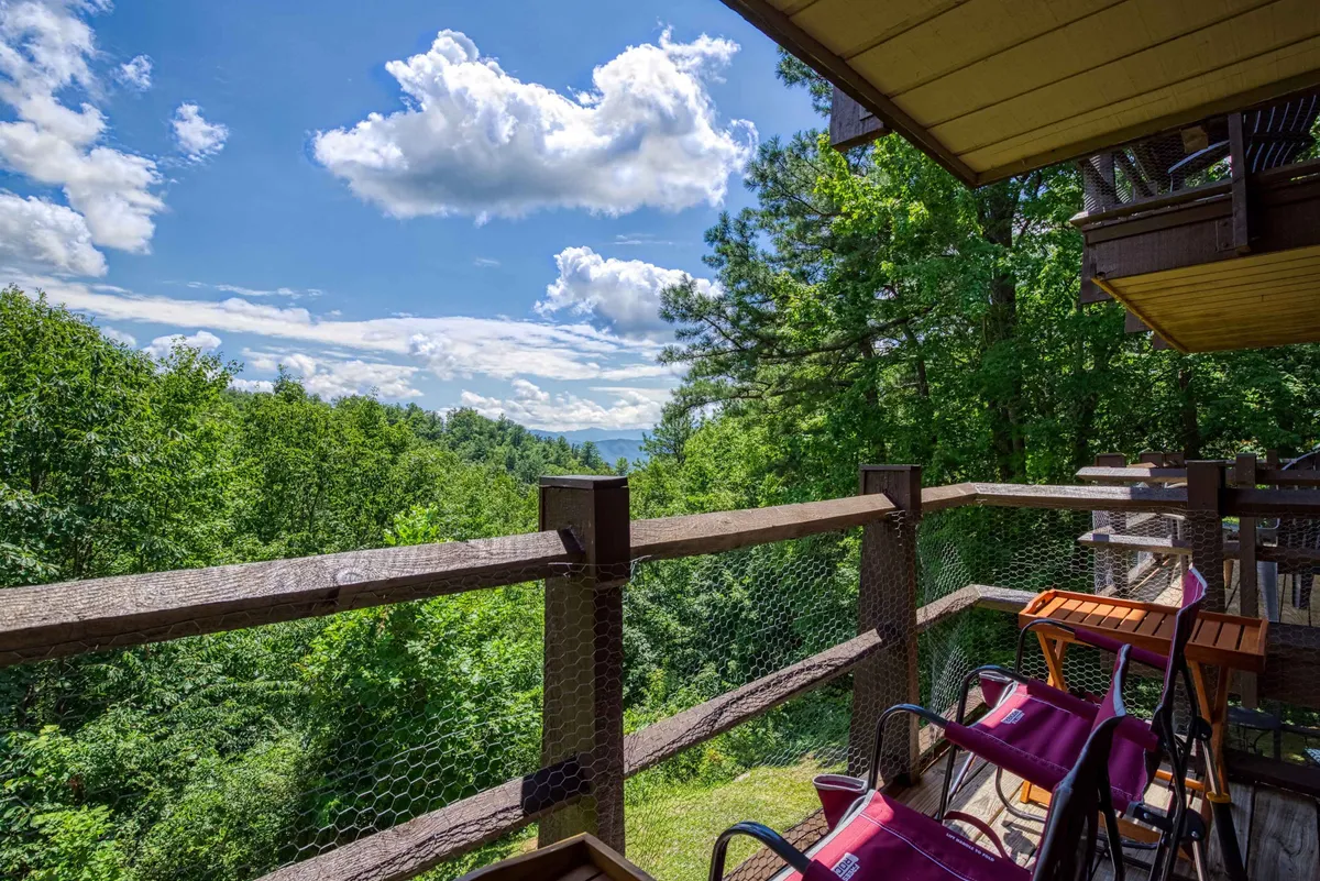 This image showcases a wooden deck with comfortable chairs, overlooking a stunning mountain view beneath a cloudy sky. The lush green trees add a sense of seclusion, while the expansive vista invites guests to enjoy tranquil sunrises or peaceful moments of relaxation. The deck's setup hints at an intimate space ideal for couples to share coffee or unwind together.