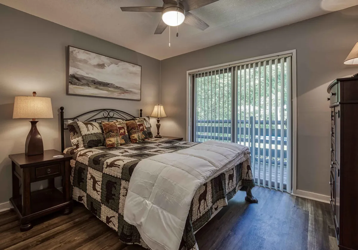 This image features a cozy bedroom with a bed dressed in a patterned quilt, wooden nightstands on each side, and a large window with blinds allowing natural light to softly illuminate the space. The décor is warm and inviting, creating an atmosphere of comfort and peace.