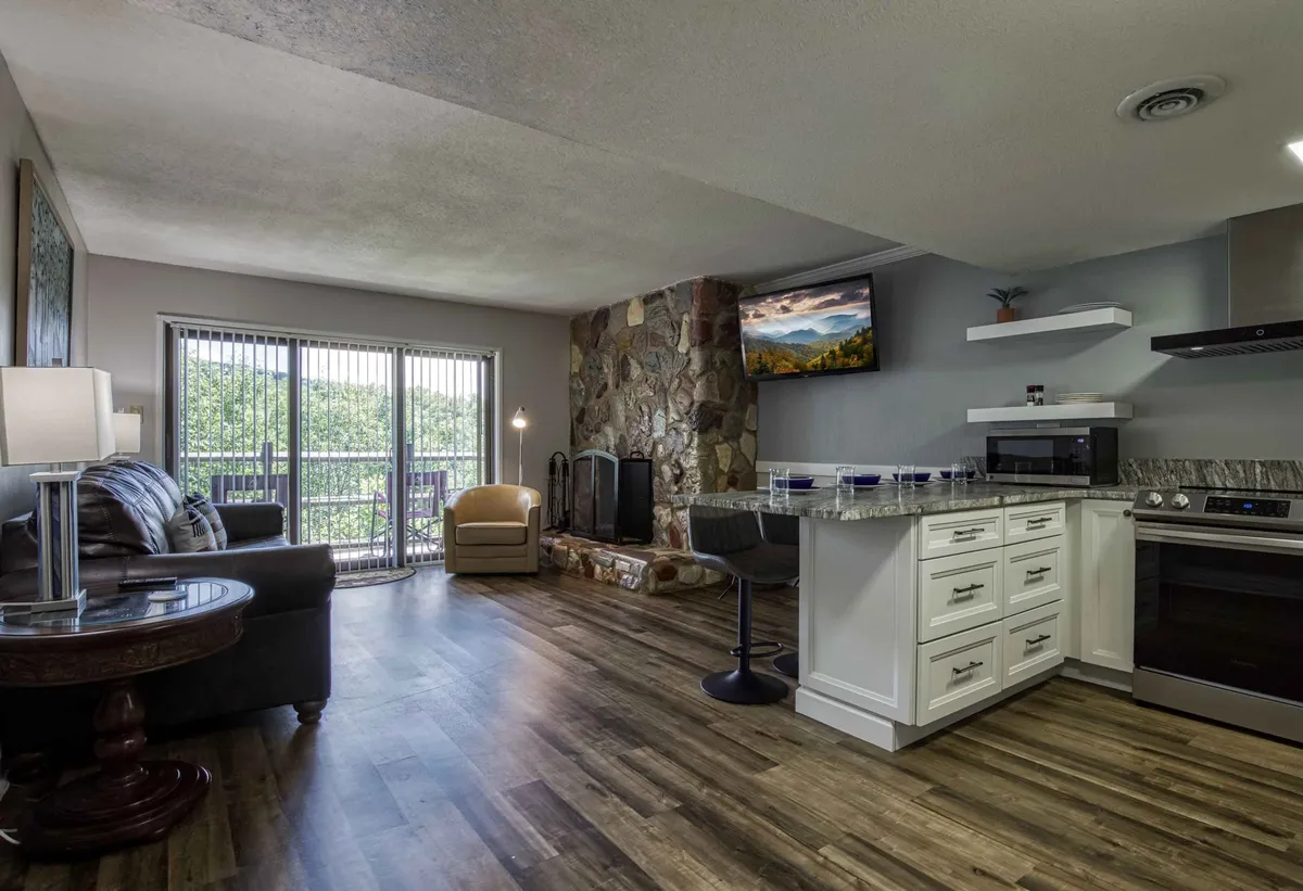 A spacious open-plan living area featuring a cozy stone fireplace, comfortable seating, and large windows that let in abundant natural light. Modern kitchen appliances and stylish furnishings complete the inviting setting, while the windows frame sweeping views of the Smoky Mountains.