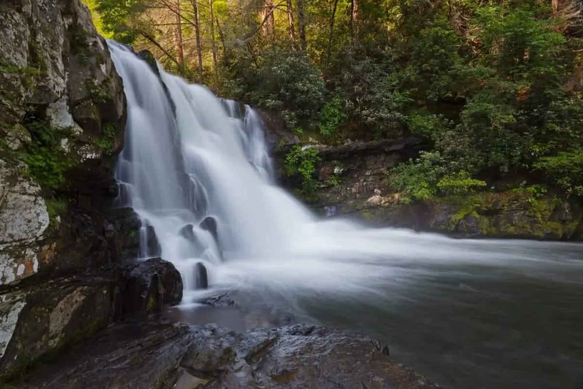 A serene waterfall pours over rocks into a calm pool, bordered by lush greenery and shaded by trees. The tranquil setting invites exploration and relaxation, resonating with the natural beauty and outdoor experiences described in the section.