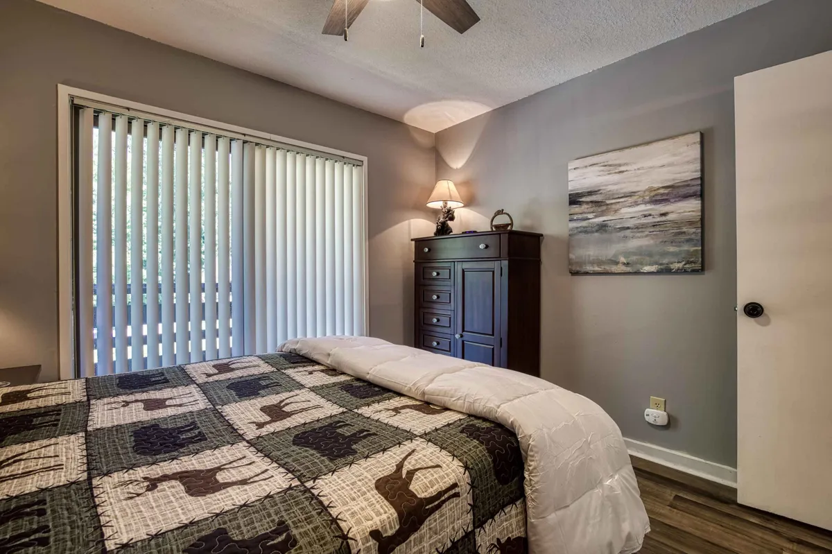 A serene bedroom with a quilted coverlet on the bed, a dresser topped with a lamp, and a window with vertical blinds. The scene is uncluttered and peaceful, ideal for restful sleep or quiet reading moments.