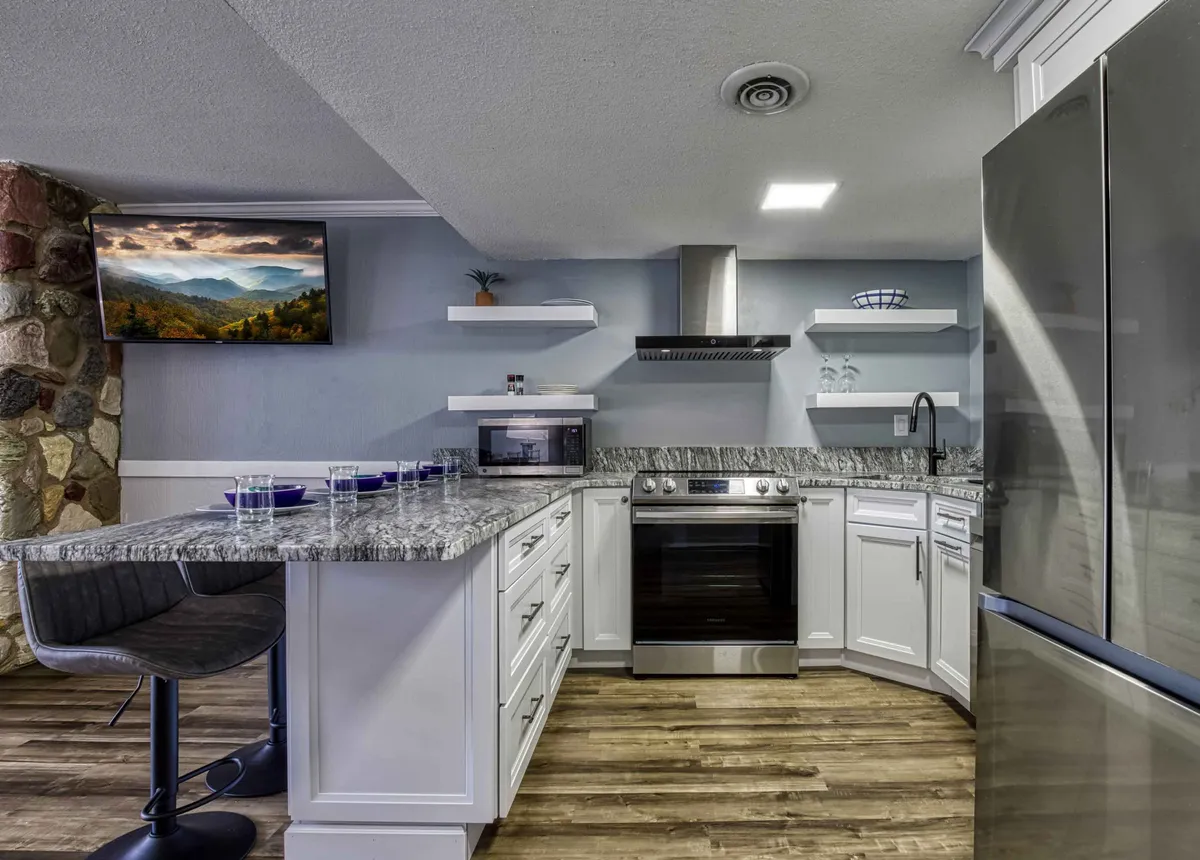 A welcoming interior scene showing a modern kitchen and part of the living area, anchored by a granite countertop and stainless steel appliances. A television is positioned so that mountain scenery is visible through adjacent windows, reinforcing the connection between interior comfort and the outdoors.